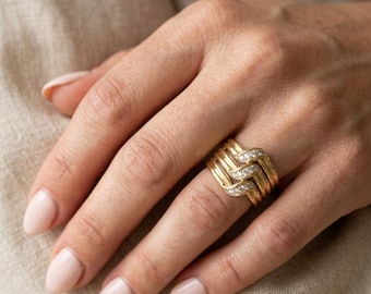 May include: A wide gold band ring featuring a woven or interlocking design. The ring consists of multiple polished gold strands, with one central strand embedded with a row of small, clear, round-cut stones that sparkle against the metal.