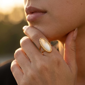 Puede incluir: Un anillo de tono dorado que presenta una gran incrustación ovalada y lisa de nácar blanco engastada en un marco de metal fino. La banda del anillo se divide en dos hilos delicados y paralelos de color dorado que conectan con el engaste central.