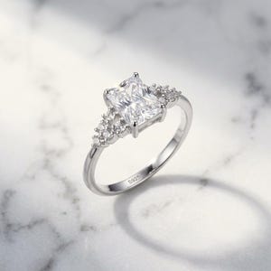 May include: A silver-toned ring featuring a large rectangular radiant-cut clear center stone flanked by a cluster of three smaller round clear stones on each side. The inner band is clearly engraved with the hallmark S925, indicating sterling silver.
