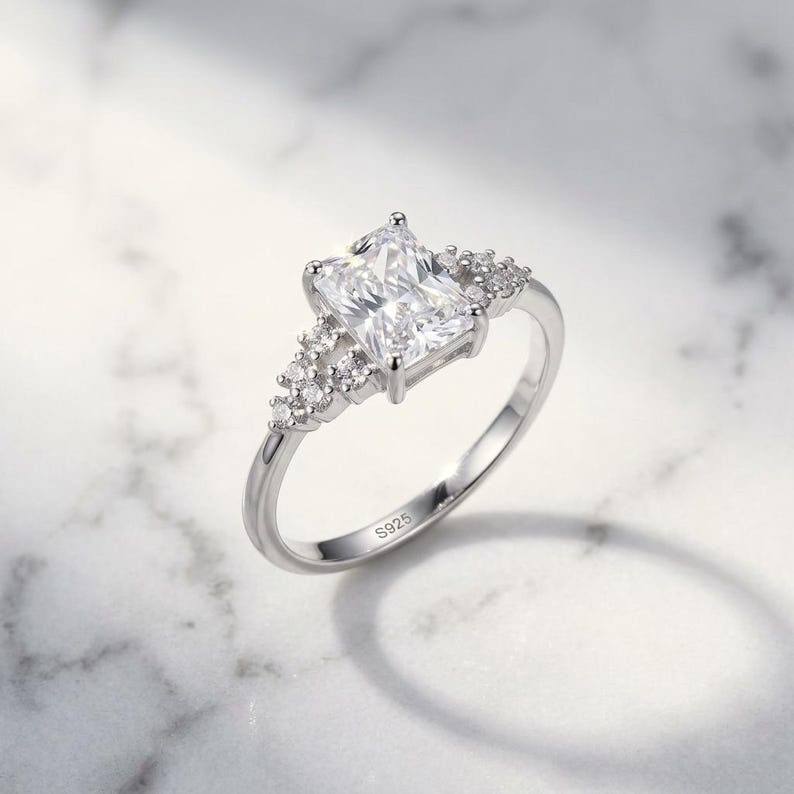 May include: A silver-toned ring featuring a large rectangular radiant-cut clear center stone flanked by a cluster of three smaller round clear stones on each side. The inner band is clearly engraved with the hallmark S925, indicating sterling silver.