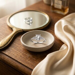 May include: A silver-toned ring featuring a central rectangular-cut clear stone flanked by smaller round stones on each side. The ring is positioned inside a small, round, beige ceramic dish, with a reflection visible in a nearby mirror.