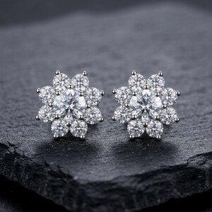 Puede incluir: Un par de pendientes de botón plateados, cada uno con una piedra preciosa central redonda y transparente rodeada por un halo de ocho piedras más pequeñas, creando un diseño floral con un acabado brillante y altamente reflectante.
