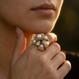 May include: A gold ring featuring a cluster of round white pearls and small pavé-set clear stones. The design consists of multiple pearl spheres arranged in a floral-like formation, with the metal band encrusted with tiny sparkling stones.