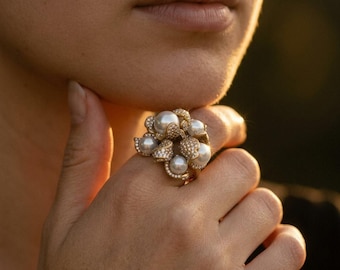 Puede incluir: Un anillo de oro con un grupo de perlas blancas redondas y pequeñas piedras transparentes engastadas en pavé. El diseño consiste en múltiples esferas de perlas dispuestas en una formación floral, con la banda de metal incrustada de diminutas piedras brillantes.