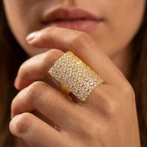 Könnte beinhalten: Ein breiter goldfarbener Ring mit einer rechteckigen Vorderseite, die vollständig mit zahlreichen kleinen, klaren, funkelnden Steinen in einem Gittermuster bedeckt ist. Die Ringschiene ist glatt und metallisch und bildet einen Kontrast zur texturierten Oberfläche.