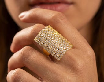 Puede incluir: Un anillo ancho de tono dorado con una cara rectangular completamente cubierta por numerosas piedras pequeñas, claras y brillantes dispuestas en un patrón de cuadrícula. La banda es lisa y metálica, contrastando con la superficie texturizada.
