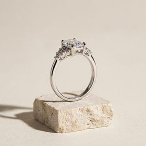 May include: A silver-toned ring featuring a large rectangular center stone held by four prongs, flanked by a cluster of three smaller round stones on each side. The inner band is clearly engraved with the text S925, indicating the metal composition.