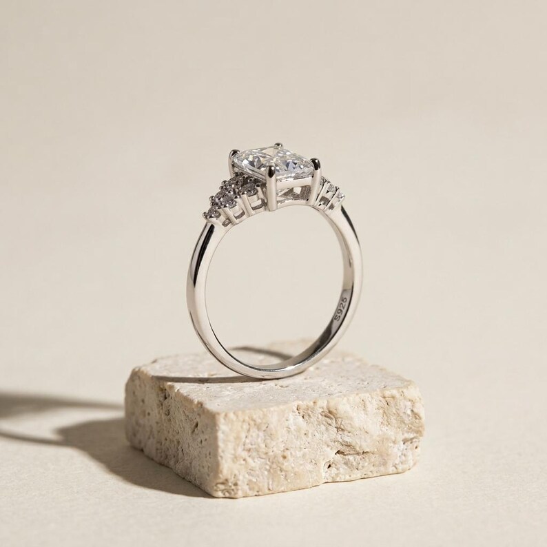 May include: A silver-toned ring featuring a large rectangular center stone held by four prongs, flanked by a cluster of three smaller round stones on each side. The inner band is clearly engraved with the text S925, indicating the metal composition.