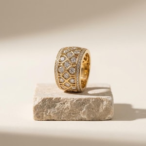 May include: A wide gold band ring featuring a central lattice pattern filled with round brilliant cut diamonds. The edges of the ring are lined with smaller pavé set diamonds, creating a textured, reflective surface across the entire gold band.