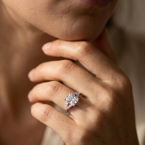May include: A silver-toned ring featuring a large, rectangular radiant-cut center stone flanked by smaller, round-cut accent stones set along the band. The metal band has a polished finish, and the stones are clear and faceted to reflect light.