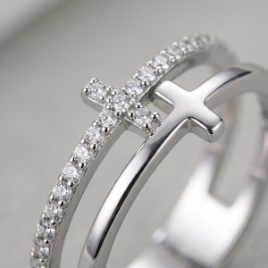 May include: A silver-toned double band ring featuring two cross motifs. One band is encrusted with small, clear, round-cut stones, while the other band is smooth and polished, each supporting a cross element of the same respective finish.