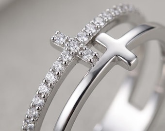 May include: A silver-toned double band ring featuring two cross motifs. One band is encrusted with small, clear, round-cut stones, while the other band is smooth and polished, each supporting a cross element of the same respective finish.