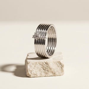Puede incluir: Un anillo de varias bandas en tono plateado que presenta cinco bandas paralelas, lisas y pulidas, unidas entre sí. Un pequeño grupo cuadrado de diminutas piedras claras y brillantes está engastado en la parte superior del anillo, añadiendo un detalle sutil y reflectante a la superficie metálica.