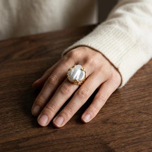 Puede incluir: Un anillo con una gran perla barroca blanca de forma irregular, engastada en un marco de metal dorado con textura orgánica que rodea la perla. El aro del anillo es fino y metálico, creando un contraste con la superficie brillante de la perla.