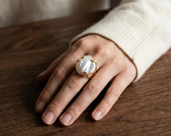 May include: A ring featuring a large, irregular white baroque pearl set within a textured, organic gold-toned metal frame that wraps around the pearl. The band of the ring is thin and metallic, contrasting with the lustrous surface of the pearl.