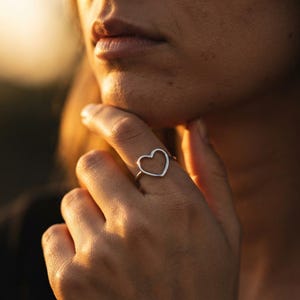 Puede incluir: Un anillo de color plateado con un diseño de corazón abierto y fino. La banda es delgada y circular, sosteniendo una pieza central en forma de corazón hueco que se asienta sobre el dedo. El metal tiene un acabado liso y pulido.