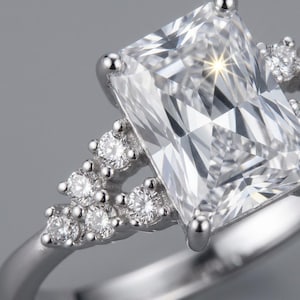 Puede incluir: Un anillo de tono plateado con una gran piedra central rectangular de facetas transparentes sujeta por cuatro garras. La banda está adornada a cada lado con un grupo de tres piedras más pequeñas, redondas y facetadas en triángulo.