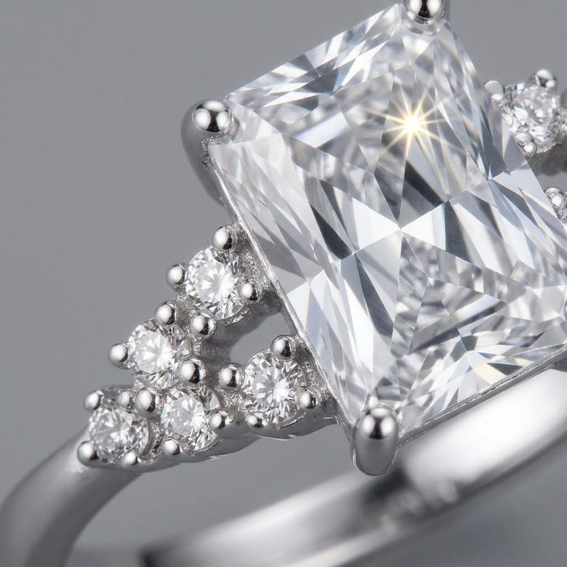 May include: A silver-toned ring featuring a large, rectangular, faceted clear center stone held by four prongs. The band is accented on each side by a cluster of three smaller, round, faceted clear stones set in a triangular arrangement.