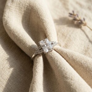 May include: A silver-toned ring featuring a large, rectangular, radiant-cut center stone. The band is accented with small, round, clear stones set along the shoulders, creating a sparkling, multi-stone design that catches the light.