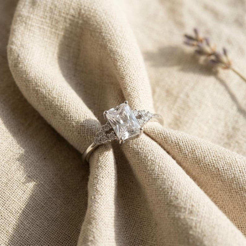May include: A silver-toned ring featuring a large, rectangular, radiant-cut center stone. The band is accented with small, round, clear stones set along the shoulders, creating a sparkling, multi-stone design that catches the light.
