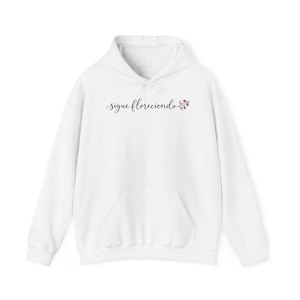May include: A white hooded sweatshirt featuring a front kangaroo pocket and long sleeves. The chest displays the black cursive text "sigue floreciendo" followed by a small illustration of a floral sprig with red and green details.