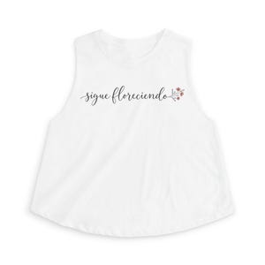 May include: A white sleeveless tank top featuring the black cursive text "sigue floreciendo" printed across the chest, accompanied by a small illustration of a branch with tiny pink flowers at the end of the lettering.