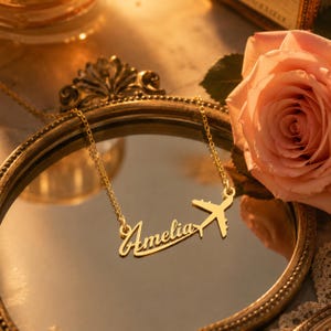 以下が含まれることがあります： 筆記体で「Amelia」と刻まれたゴールドトーンのメタル製ネームネックレス。名前の最後には小さな飛行機のシルエットが付いています。ネックレスは、アンティーク調の装飾が施された円形の鏡のフレームの上に置かれています。