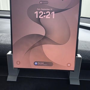 May include: A rectangular smartphone with a large, unfolded screen displaying a lock screen with the time 12:21, the date Sun, September 13, and a soft pink abstract wave background. The device is held upright by a light grey, 3D-printed plastic stand.