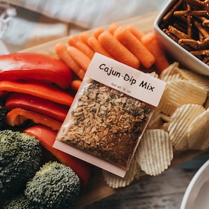 May include: A wooden serving board with a variety of snacks, including broccoli florets, red bell pepper slices, baby carrots, potato chips, and pretzel sticks. A small pink packet labeled "Cajun-Dip Mix, Net wt. 1.5 oz" is in the center of the board.