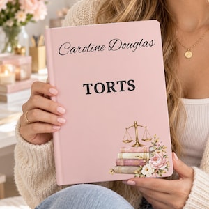 May include: A light pink hardcover notebook featuring the name Caroline Douglas in black script, the word TORTS in bold black capital letters, and a graphic of a gold scale of justice resting on a stack of books with pink and white flowers.