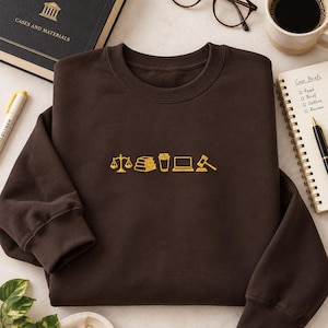 May include: A dark brown crewneck sweatshirt featuring a centered yellow embroidered graphic. The design consists of a series of five icons: a scale of justice, a stack of books, a coffee cup, a laptop computer, and a wooden judge's gavel.