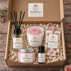 May include: A gift box containing a reed diffuser with black sticks, a candle with a pink rose-shaped wax top, a wax melt with dried flowers, a jar of pink sugar scrub, and a small bottle of essential oil, all labeled Petal & Grove Haus.