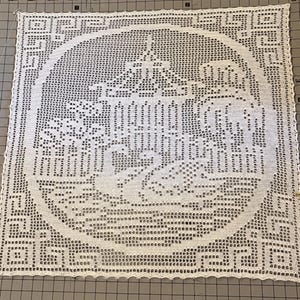 May include: A square white crochet doily featuring a central circular motif with a detailed scene of a swan swimming near a gazebo and trees. The border is decorated with a geometric Greek key pattern, all crafted with fine white thread.
