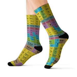 Puede incluir: Un par de calcetines con un patrón repetitivo de la tabla periódica de los elementos en tonos amarillo, azul, verde y morado. Los calcetines tienen talones y puntas negros, con símbolos químicos y números atómicos claramente visibles impresos.