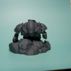 May include: A dark gray, unpainted resin miniature figurine of a heavily armored warrior in a crouching stance. The figure features detailed shoulder pauldrons and chest plating, positioned atop a base sculpted to resemble jagged, uneven rock formations.
