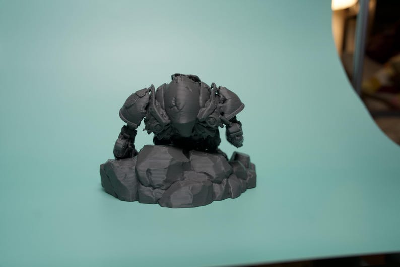 May include: A dark gray, unpainted resin miniature figurine of a heavily armored warrior in a crouching stance. The figure features detailed shoulder pauldrons and chest plating, positioned atop a base sculpted to resemble jagged, uneven rock formations.