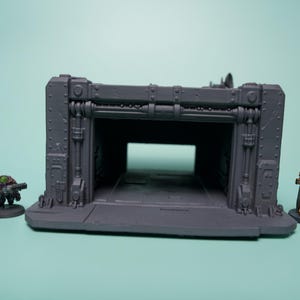 May include: A gray, unpainted industrial-style miniature terrain structure featuring a wide, open rectangular bay with reinforced paneling and rivets. Flanking the structure are two small painted gaming miniatures, one in dark tones and one in gold.