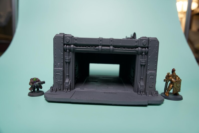 May include: A gray, unpainted industrial-style miniature terrain structure featuring a wide, open rectangular bay with reinforced paneling and rivets. Flanking the structure are two small painted gaming miniatures, one in dark tones and one in gold.