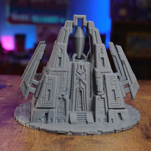 May include: A gray 3D printed miniature structure featuring a central spire, geometric wall carvings, and a stepped entrance. The object is set on a circular base with a textured surface, displaying intricate architectural details and sharp angles.