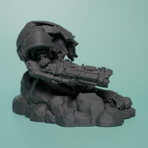 May include: A dark gray unpainted resin miniature of a mechanical warrior torso with a large mounted cannon arm. The figure is depicted in a low-profile pose, resting on a textured, rocky base with visible mechanical joints and armor plating.