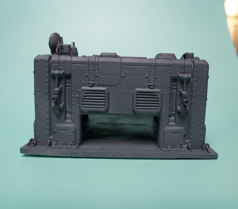 May include: A dark gray 3D printed miniature structure featuring a rectangular base, vertical wall panels with rivets, two rectangular vents, and a small satellite dish mounted on the top left corner. The structure has a hollow arched opening.