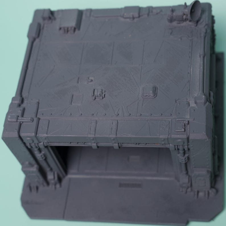 May include: A gray, unpainted plastic miniature terrain piece featuring a square, bunker-like structure with a flat roof, reinforced corner pillars, and a wide, open archway entrance. The surface is detailed with molded panel lines and a small satellite dish.