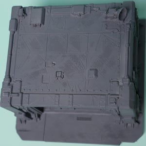 May include: A gray, unpainted plastic miniature terrain piece featuring a rectangular structure with a flat roof. The surface is detailed with etched geometric panel lines, small square vents, and a circular pipe fitting on the top corner.