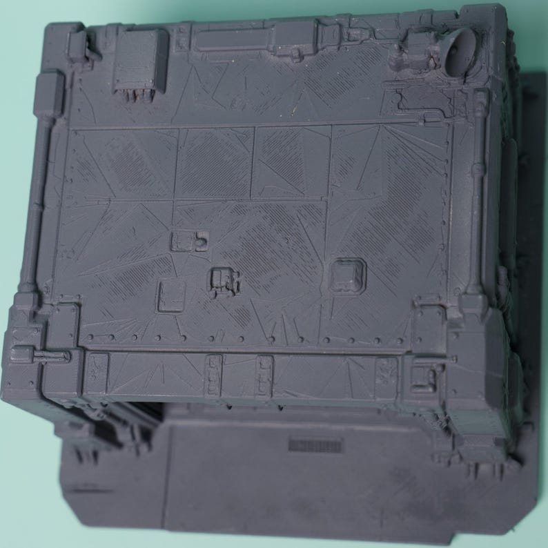 May include: A gray, unpainted plastic miniature terrain piece featuring a rectangular structure with a flat roof. The surface is detailed with etched geometric panel lines, small square vents, and a circular pipe fitting on the top corner.