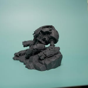 May include: A dark gray, unpainted resin miniature of a futuristic mech warrior in a kneeling position. The figure features mechanical armor plating, a rounded helmet, and integrated weaponry, all mounted on a textured, rocky terrain base.