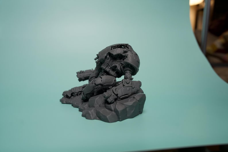 May include: A dark gray, unpainted resin miniature of a futuristic mech warrior in a kneeling position. The figure features mechanical armor plating, a rounded helmet, and integrated weaponry, all mounted on a textured, rocky terrain base.