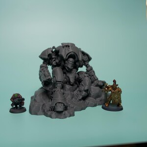 May include: A gray unpainted resin miniature of a large armored robot sitting on a rocky base, flanked by a small green-clad figure on the left and a smaller gold-painted armored figure holding a shield and spear on the right side.