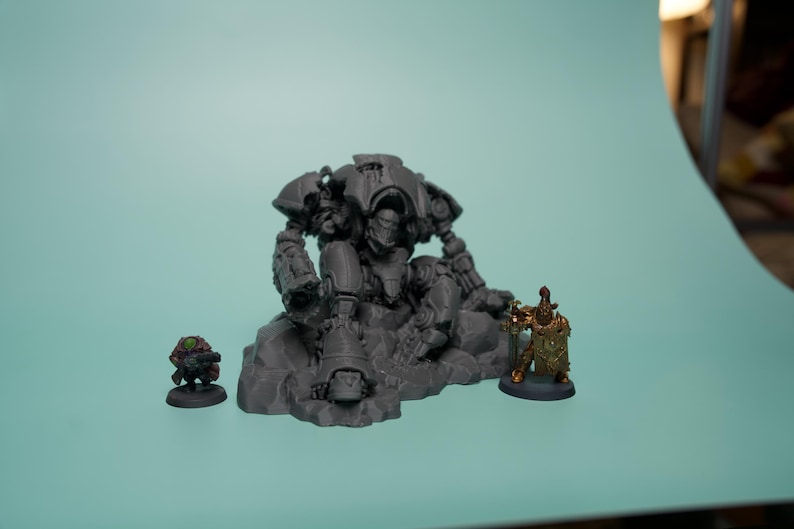 May include: A gray unpainted resin miniature of a large armored robot sitting on a rocky base, flanked by a small green-clad figure on the left and a smaller gold-painted armored figure holding a shield and spear on the right side.