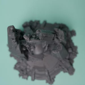 Może przedstawiać: Ciemnoszara, niepomalowana figurka miniaturowa o złożonej, poszarpanej strukturze z wieloma wystającymi elementami geometrycznymi i ostrymi, kanciastymi powierzchniami. Obiekt posiada okrągłą podstawę oraz gęstą, teksturowaną kompozycję o wielu poziomach.