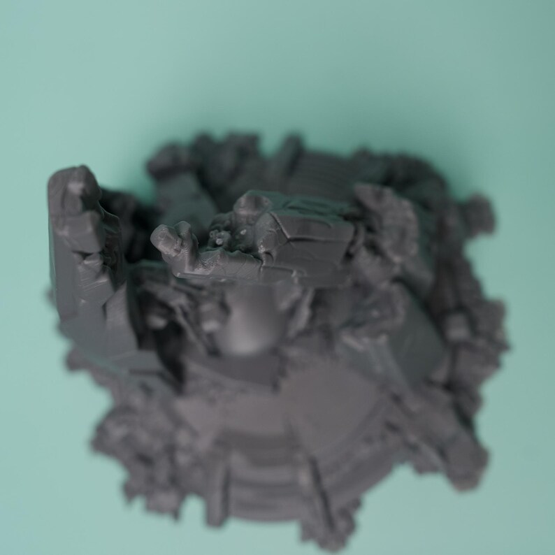 Może przedstawiać: Ciemnoszara, niepomalowana figurka miniaturowa o złożonej, poszarpanej strukturze z wieloma wystającymi elementami geometrycznymi i ostrymi, kanciastymi powierzchniami. Obiekt posiada okrągłą podstawę oraz gęstą, teksturowaną kompozycję o wielu poziomach.